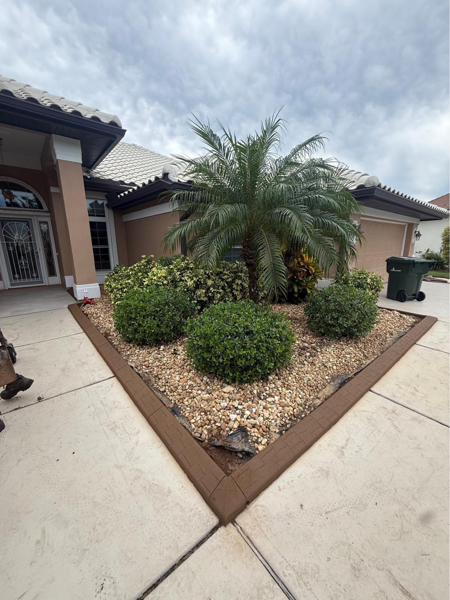 Brown curbing framing a gravel bed with trimmed shrubs and a palm at a driveway